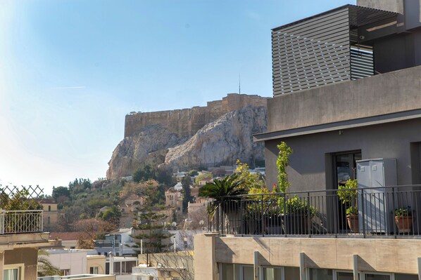 A modern building with a balcony, potted plants, and a view of a rocky hill with a castle-like structure in the background. - NOOS Acropolis (Athens)