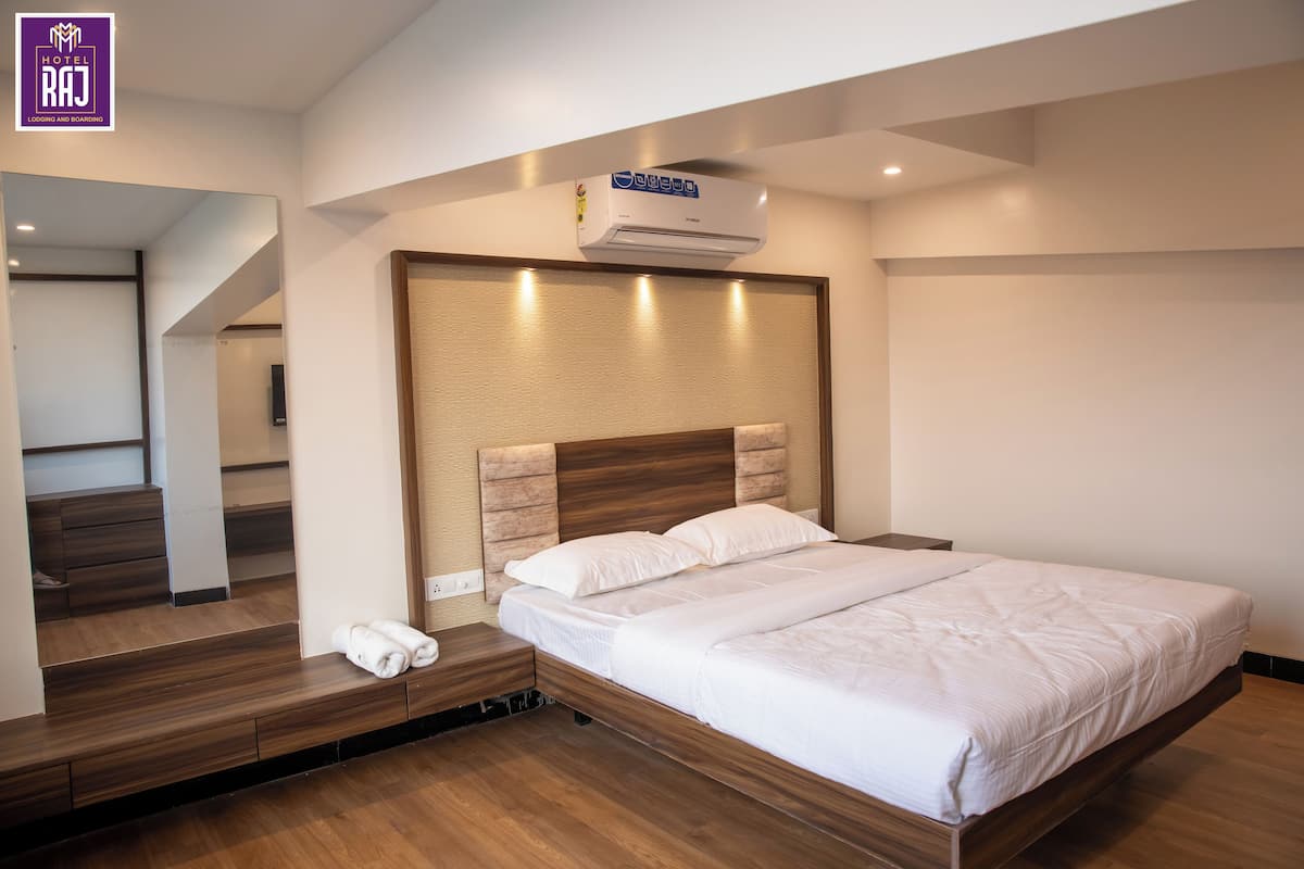 A modern hotel room with a wooden bed, a built-in bench with towels, a wall-mounted air conditioner, and a mirror reflecting the room.