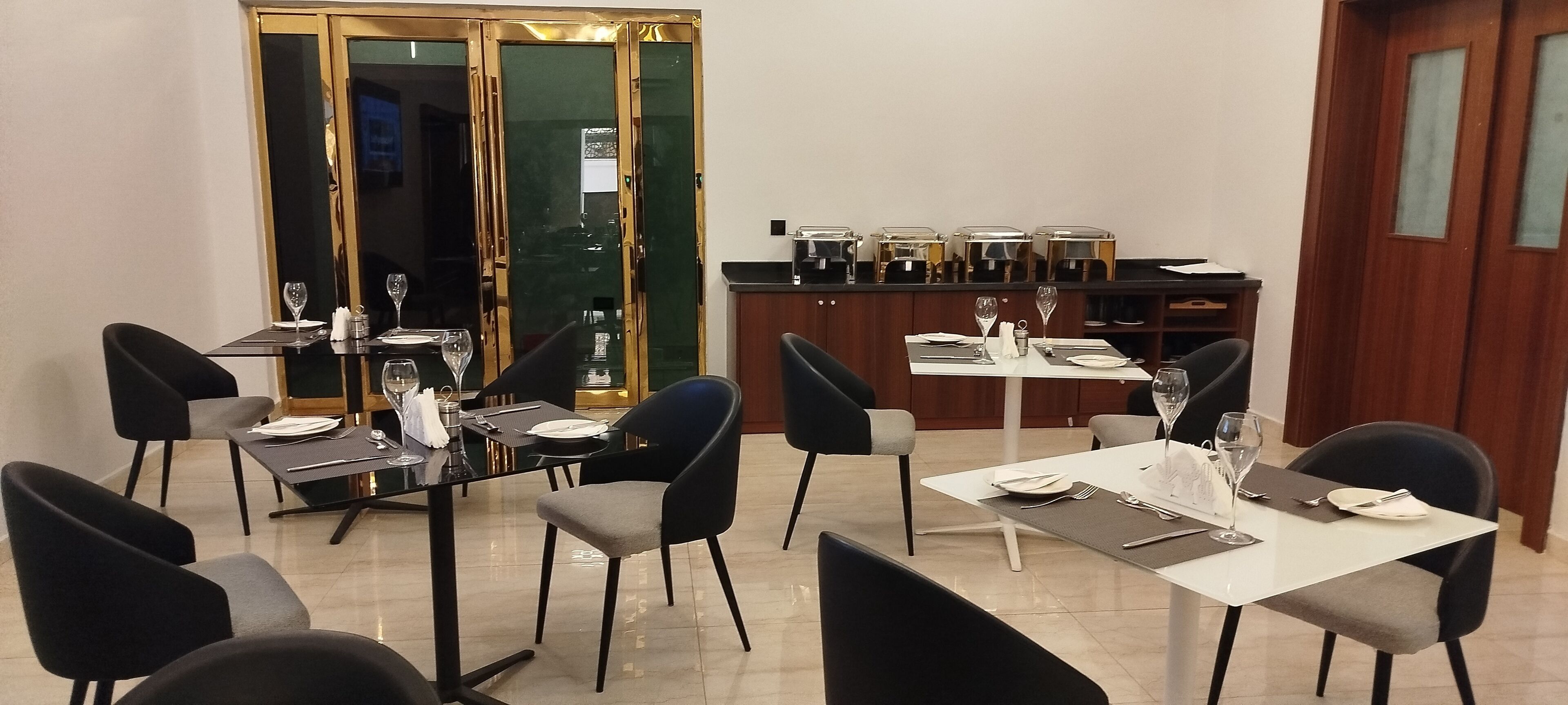 A dining area with tables set for two, black chairs, and a buffet line with food containers.