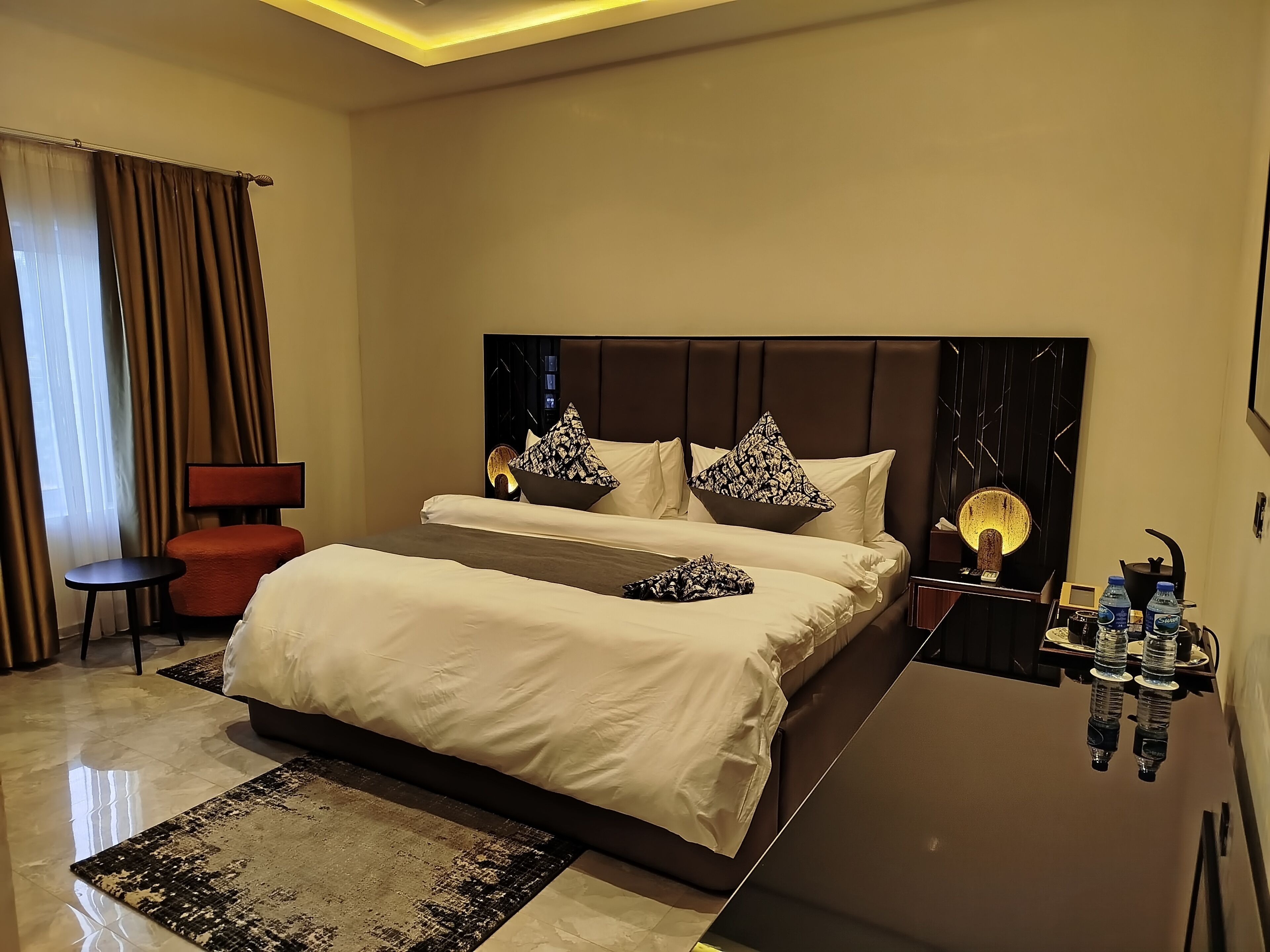 A hotel room with a large bed, a bedside table with a lamp, a red chair, and a minibar.