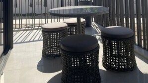 A balcony with a round glass table and two wicker stools, surrounded by a metal fence.