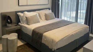 A modern bedroom with a large bed, a bedside table with a lamp, and a view of the outdoors through large windows.