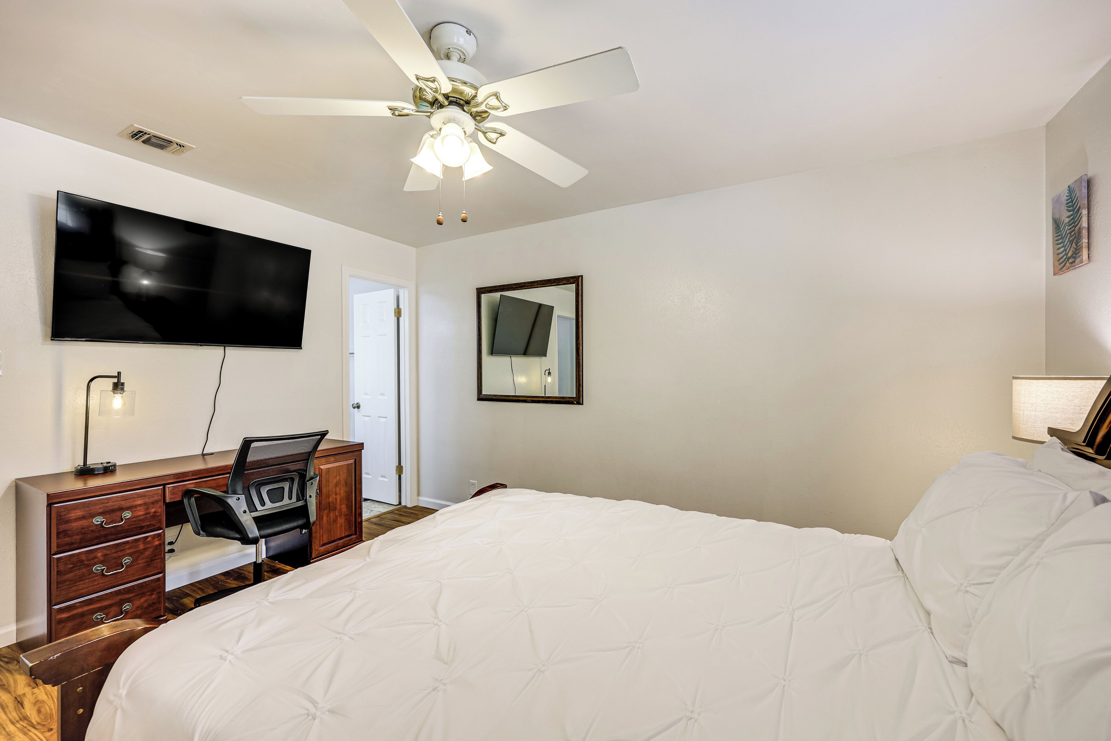 Une chambre à coucher comprenant un lit, un bureau, une chaise, une télévision, un ventilateur de plafond et un miroir.