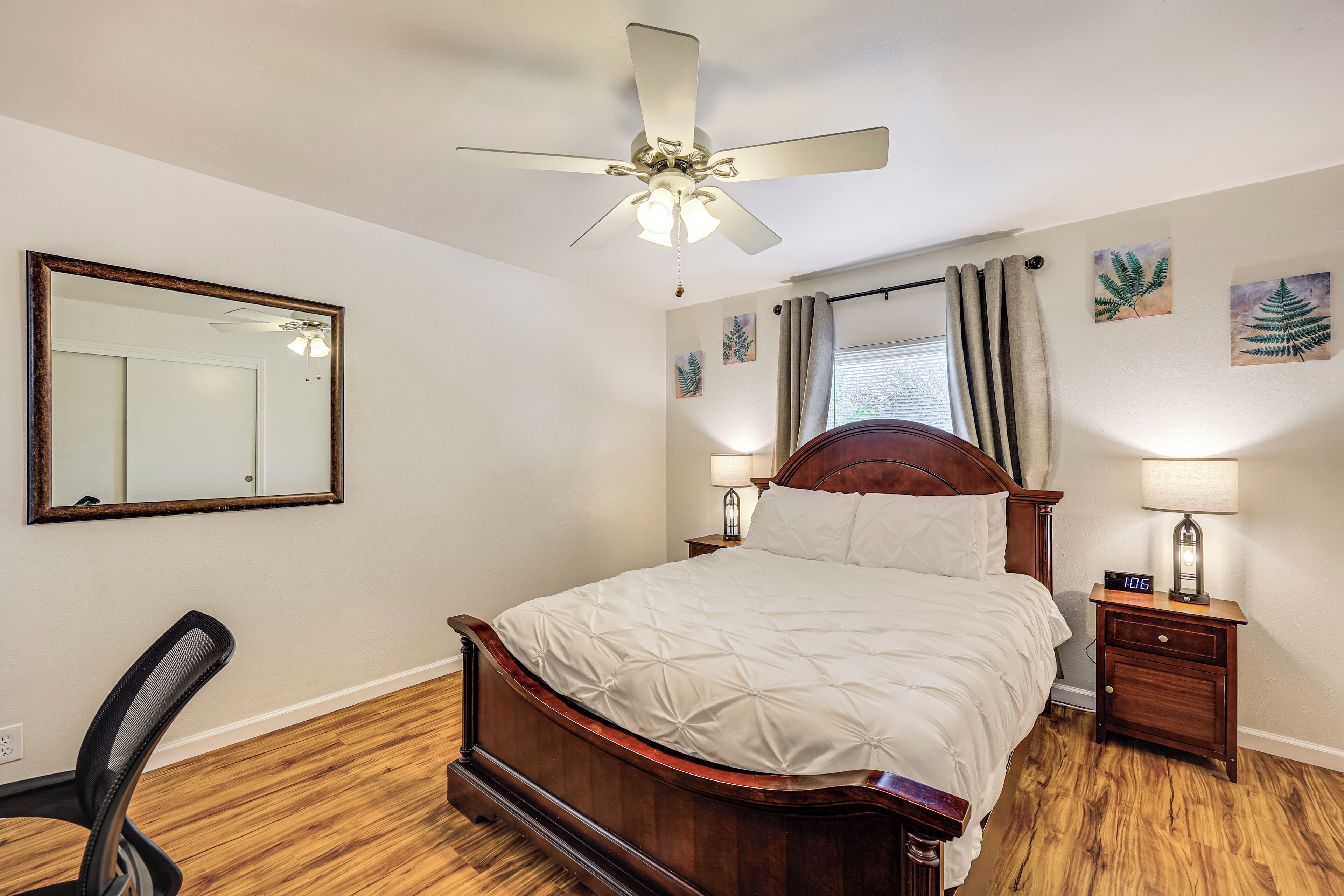 A bedroom with a bed, a ceiling fan, a nightstand with a lamp, a chair, and artwork on the walls.