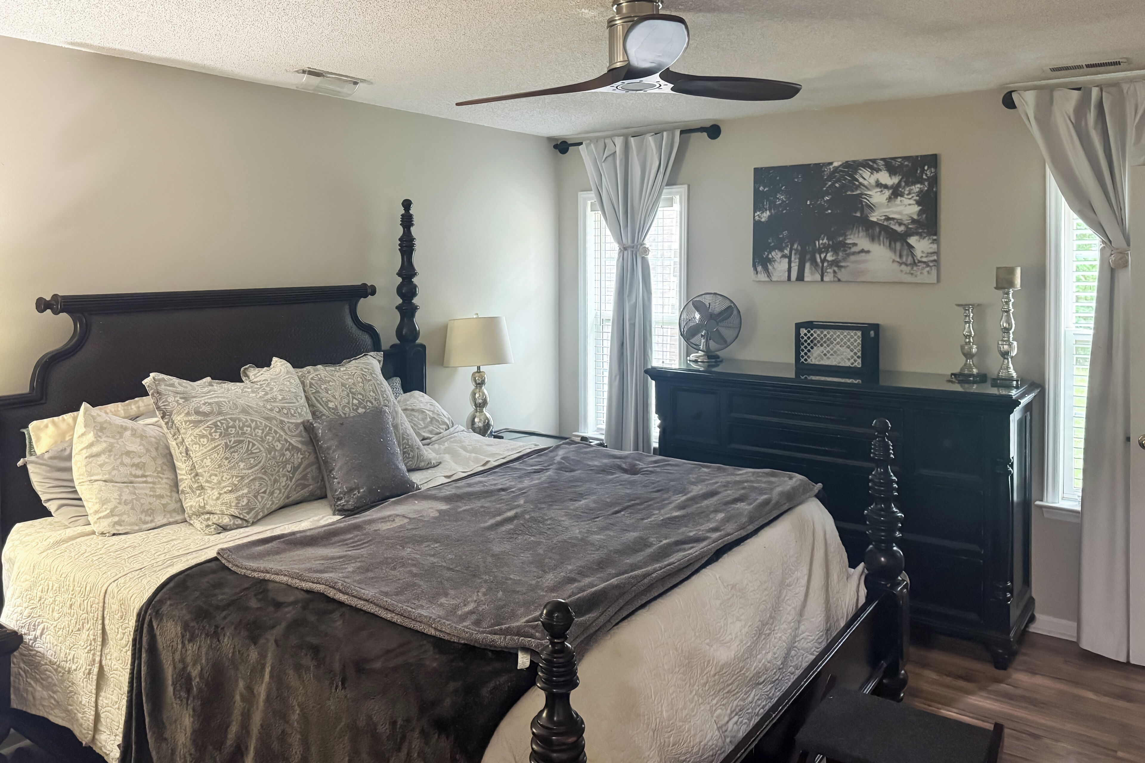A bedroom with a large bed, a ceiling fan, a dresser, and a window with curtains.