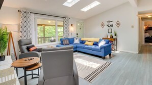 A modern living room with a sectional sofa, a grey armchair, and a small wooden table. There are skylights on the ceiling, a large window with curtains, and a wall-mounted shelf with decorative items. - Community Pools & Hot Tub: Albrightsville Cottage! (Albrightsville)
