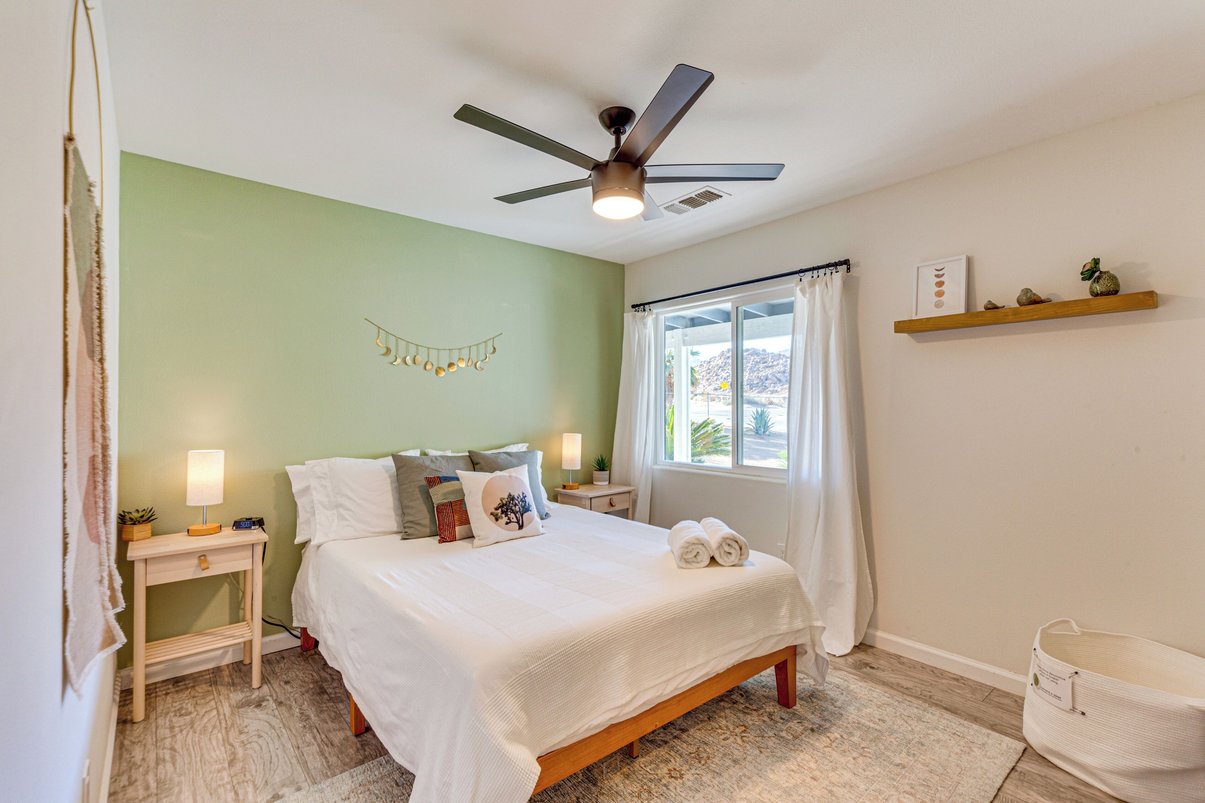 A bedroom with a bed, bedside tables, a ceiling fan, a window with curtains, and a shelf with decorative items.