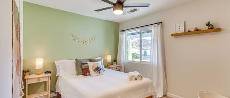 A bedroom with a bed, bedside tables, a ceiling fan, a window with curtains, and a shelf with decorative items.