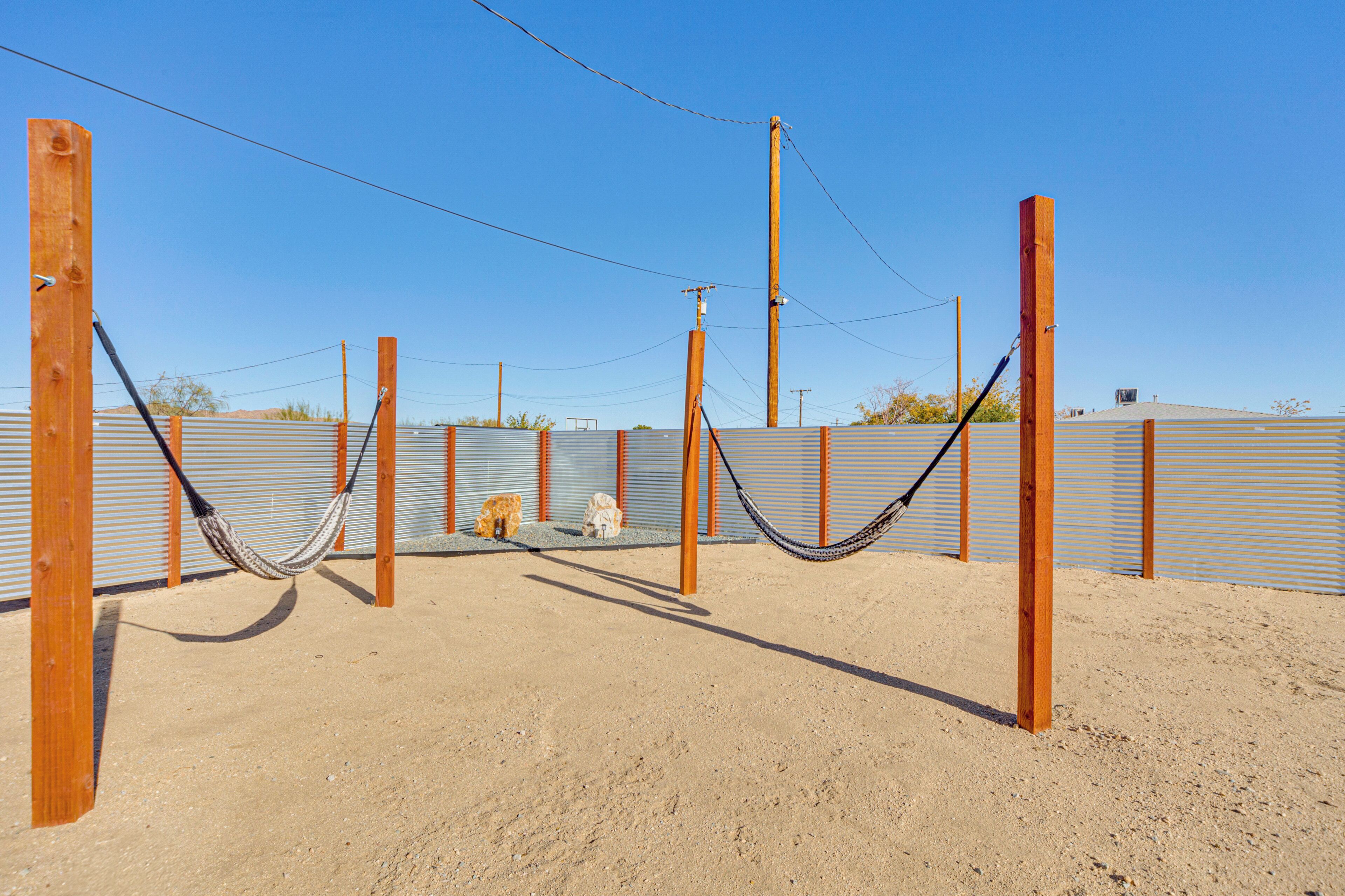 Un espacio al aire libre con arena, una hamaca entre postes de madera, una valla metálica y postes de electricidad.