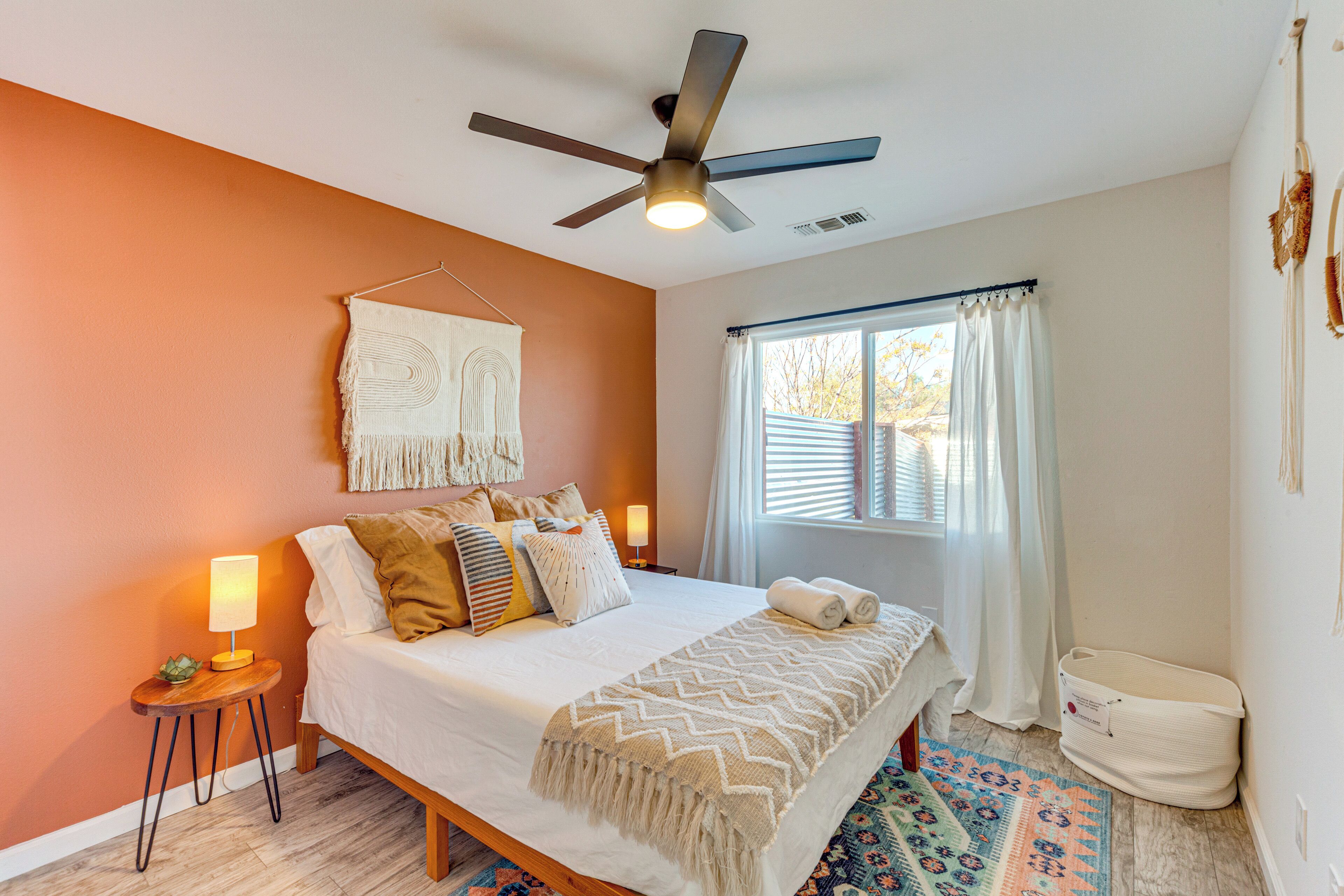 A bedroom with a bed, a ceiling fan, a bedside lamp, a window with curtains, and a small table with a lamp.