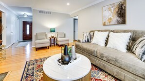 A spacious living room with a beige sofa, a round coffee table, and a large abstract painting on the wall. - Fire Pit & Yard: Family Home in Farmington Hills! (Farmington Hills)