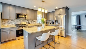A modern kitchen with grey cabinets, stainless steel appliances, a granite countertop, and two white bar stools. - Mtn-view Gem w/ Hot Tub & Fire Pit in North Conway (North Conway)