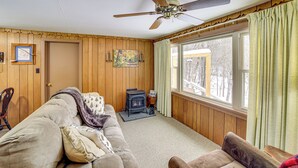 A cozy living room with a fireplace, wooden walls, a ceiling fan, and a large window with curtains. - Fish, Hike & Relax: Riverfront Manistique Cabin (Manistique)
