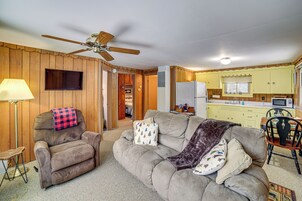 Una sala de estar con sofá, sillón y ventilador de techo. Se ve una zona de cocina al fondo.