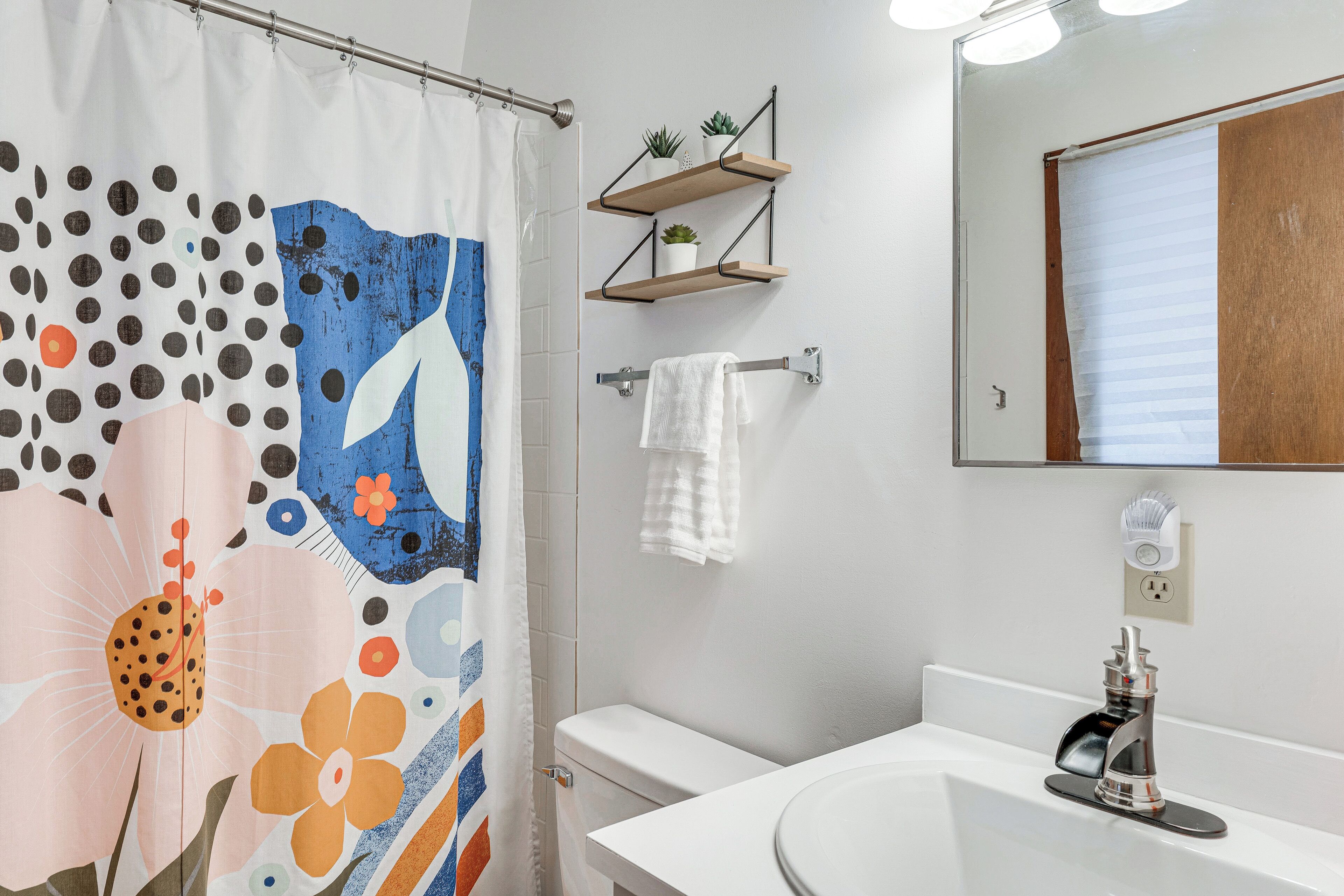 Ein Badezimmer mit Duschvorhang mit floralen und gepunkteten Mustern, einer Toilette, einem Waschbecken mit modernem Wasserhahn und Holzregalen mit Topfpflanzen.