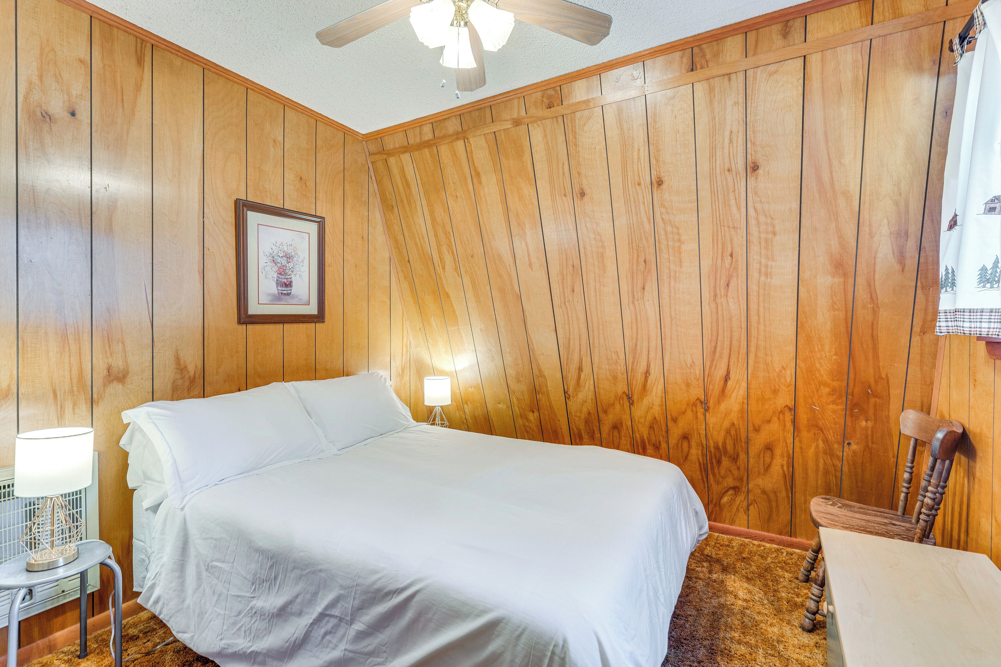 A bedroom with wooden panel walls, a bed with white bedding, a bedside lamp, a framed picture on the wall, and a ceiling fan.
