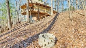 A cabin with a wooden exterior, a balcony, and a stone fire pit in a forested area. - Hot Tub, Deck & Fire Pit: Secluded Cabin in Murphy (Murphy)