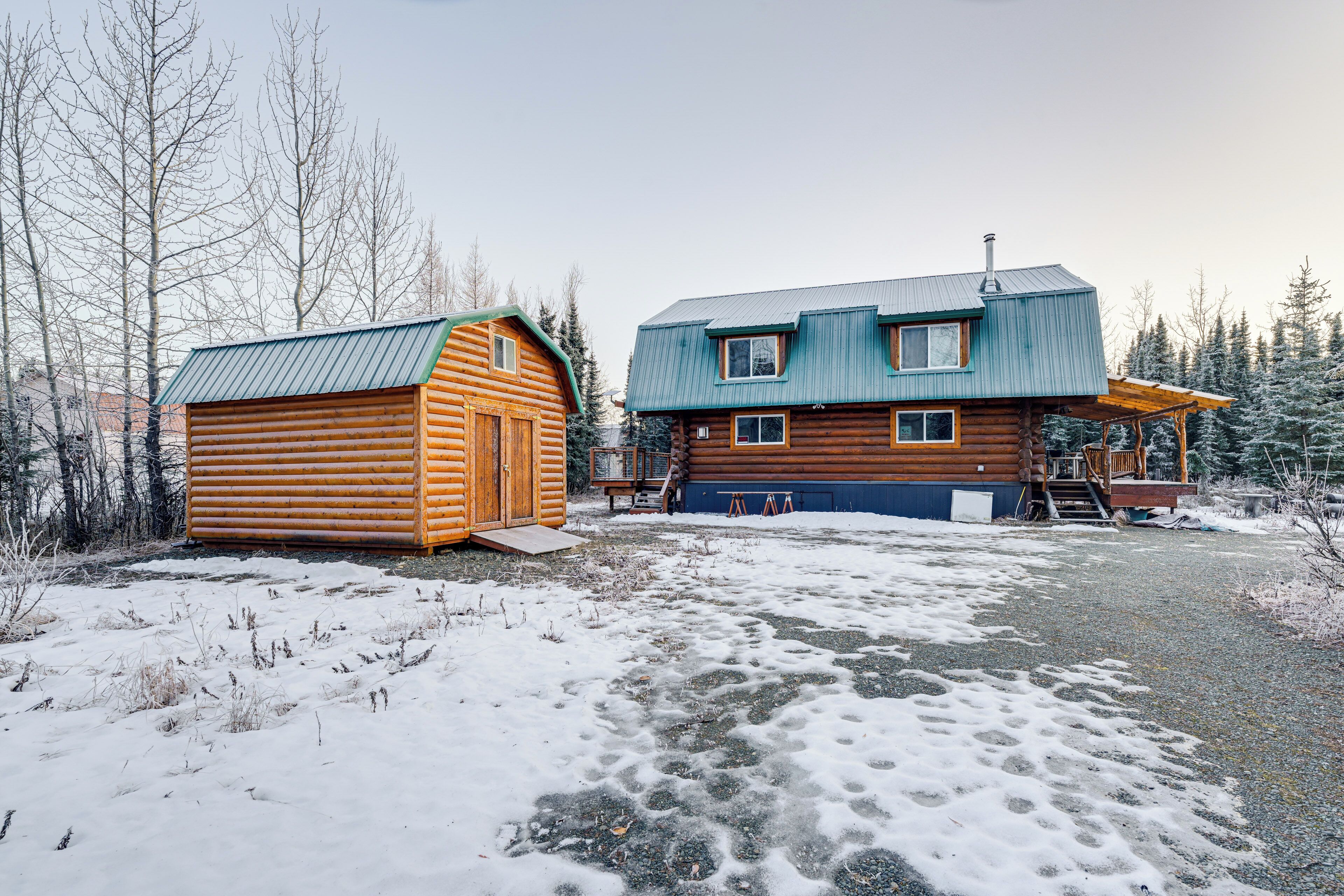 Une cabane en rondins et une maison moderne avec un toit en métal, entourées d’un paysage enneigé et d’arbres.