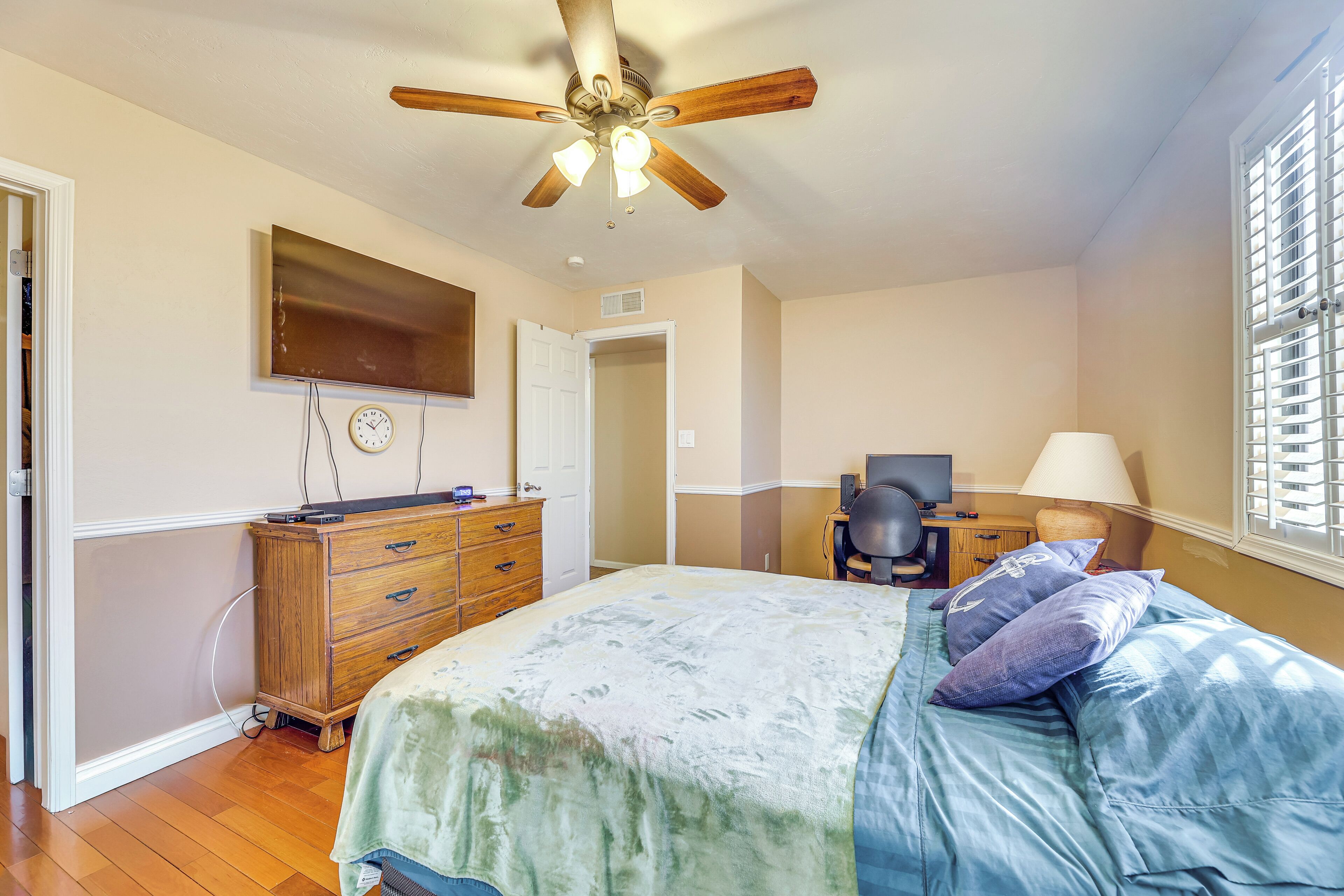 Une chambre à coucher comprenant un lit, une commode en bois, un bureau avec une chaise, un ventilateur de plafond, un téléviseur fixé au mur et une fenêtre avec des stores.
