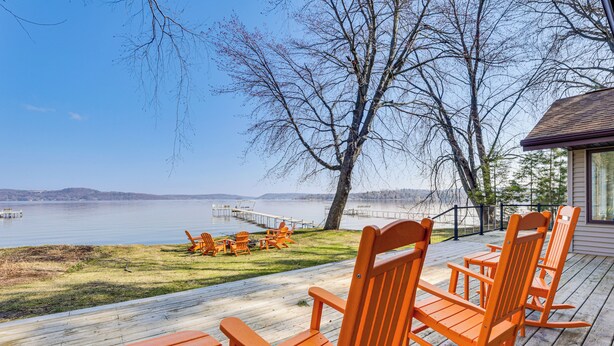 A wooden deck with orange Adirondack chairs overlooking a lake, a dock, and a building with a sloped roof. - Lake Wisconsin Retreat w/ Dock: Great for Groups! (Lodi)