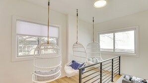 A modern interior with a wooden floor, white walls, and a staircase with a metal railing. There are three white wicker chairs hanging from the ceiling, and a sofa with cushions on the lower level. - Lake Wisconsin Retreat w/ Dock: Great for Groups! (Lodi)