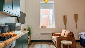 A modern kitchen with a built-in stove, a dining area with a wooden table, a sofa, and a small round table. A window with a view of a brick building is visible. - WhiskAwayStays No. 58 Studio No. 6 (Worcester)