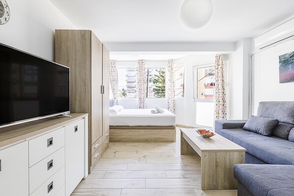 A modern apartment with a large TV, a wooden cabinet, a bed, a sofa, and a view of buildings through the window.