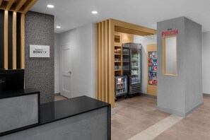 Un hall d’hôtel avec un comptoir de réception, un réfrigérateur rempli de boissons et un distributeur automatique portant l’inscription « EN CAS D’URGENCE ».