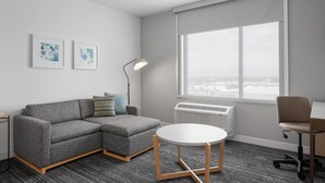 Un salon moderne avec un canapé d’angle gris, une table ronde, un lampadaire et une grande fenêtre donnant sur la ville.