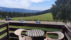 Varanda com duas cadeiras de plástico e uma mesa de madeira, com vista para um estacionamento com carros e um campo gramado com árvores.