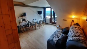 Uma sala de estar acolhedora com sofá, mesa de jantar com cadeiras, TV na parede e varanda com vista.