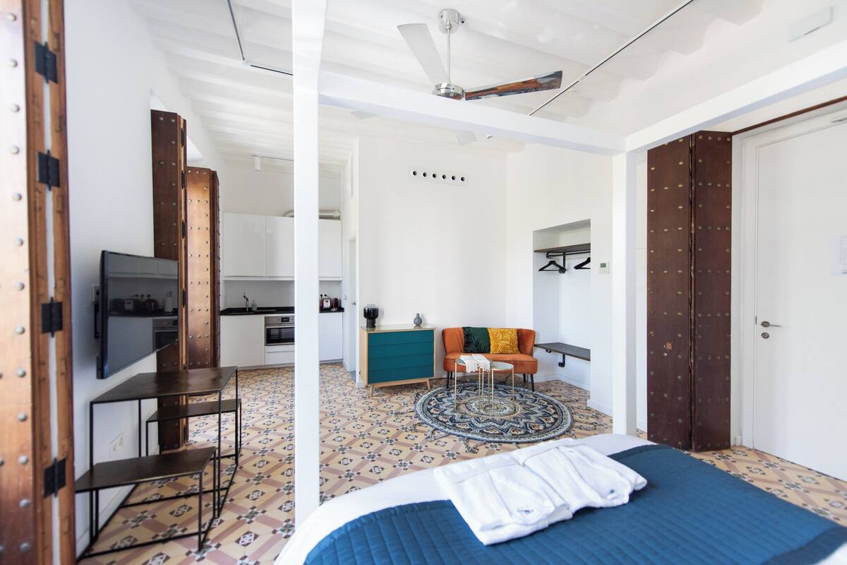 A modern hotel room with a bed, a TV, a sofa, and a kitchen area visible through an open door.