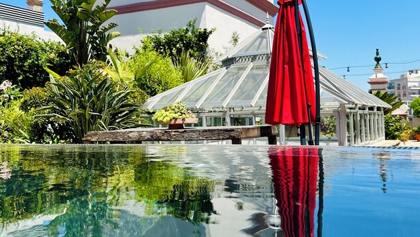 Ein klares Schwimmbecken mit einem roten Sonnenschirm, umgeben von üppiger Vegetation und einem weißen Gebäude mit Kuppeldächern.