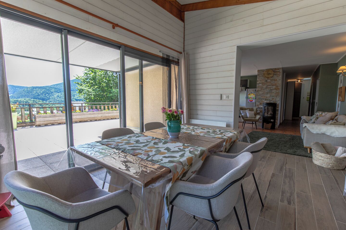 Un coin repas avec une table et des chaises, un vase rempli de fleurs et une vue sur une montagne et la verdure environnante.