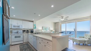Cocina moderna con armarios blancos, microondas y estufa. Hay una pequeña isla con un fregadero y una silla. La habitación tiene un balcón con vistas al océano.