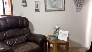 A living room with a brown leather chair, a small wooden side table with a sign, and framed botanical prints on the wall.