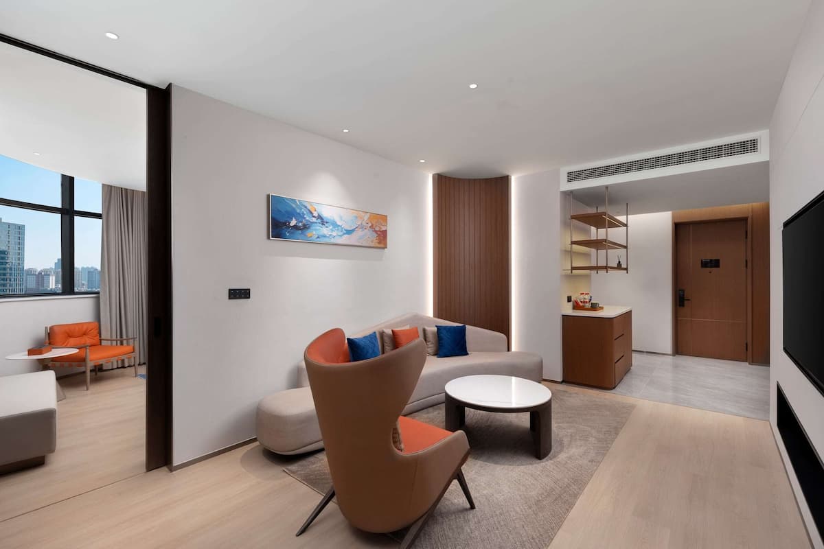 Une chambre d’hôtel moderne, équipée d’un canapé, d’un fauteuil et d’une table basse. Un tableau orne le mur, une fenêtre est habillée de rideaux et une porte donne accès à une autre pièce.