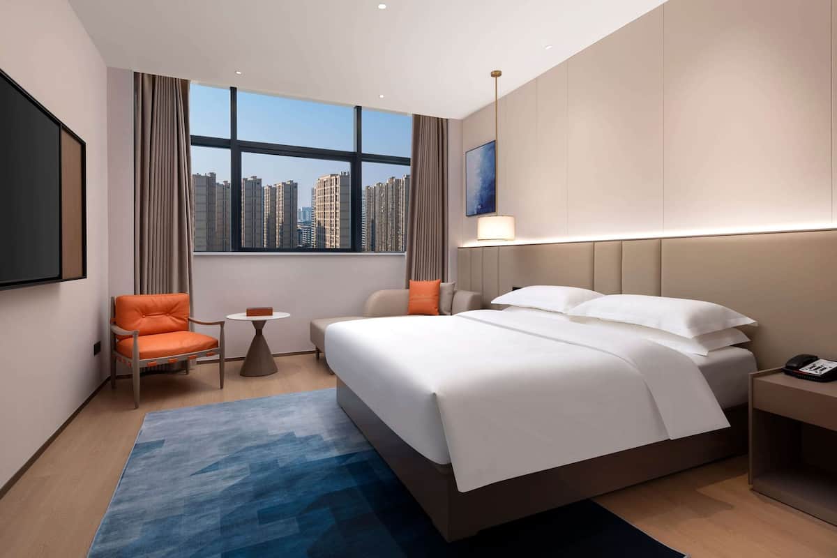 Une chambre d’hôtel moderne avec un grand lit, une petite table, une chaise et une vue sur les gratte-ciel, visible à travers une grande fenêtre.