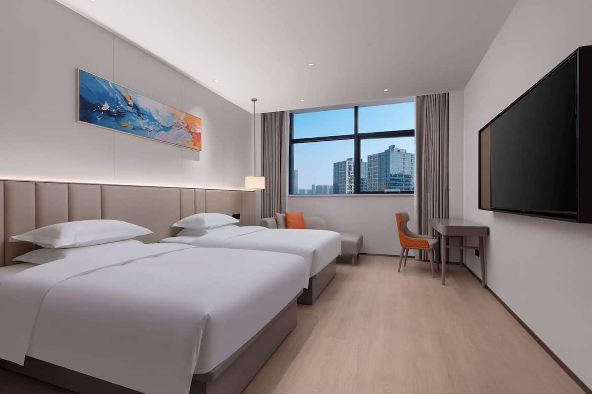 Une chambre d’hôtel avec deux lits, un bureau, une chaise, une télévision et une grande fenêtre donnant sur la ville.