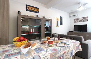 Ένας χώρος τραπεζαρίας με ένα τραπέζι στρωμένο για το γεύμα, ένα μπολ με φρούτα και μια τηλεόραση στο βάθος.