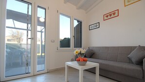 A living room with a grey sofa, a white coffee table with a bowl of fruit and flowers, and a sliding glass door leading outside. - Villetta Elena, Ac, Wifi, Lido Degli Scacchi (Comacchio)