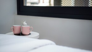 Two pink mugs on a white table with a small white figurine beside them, set against a plain wall and a window with a black frame.