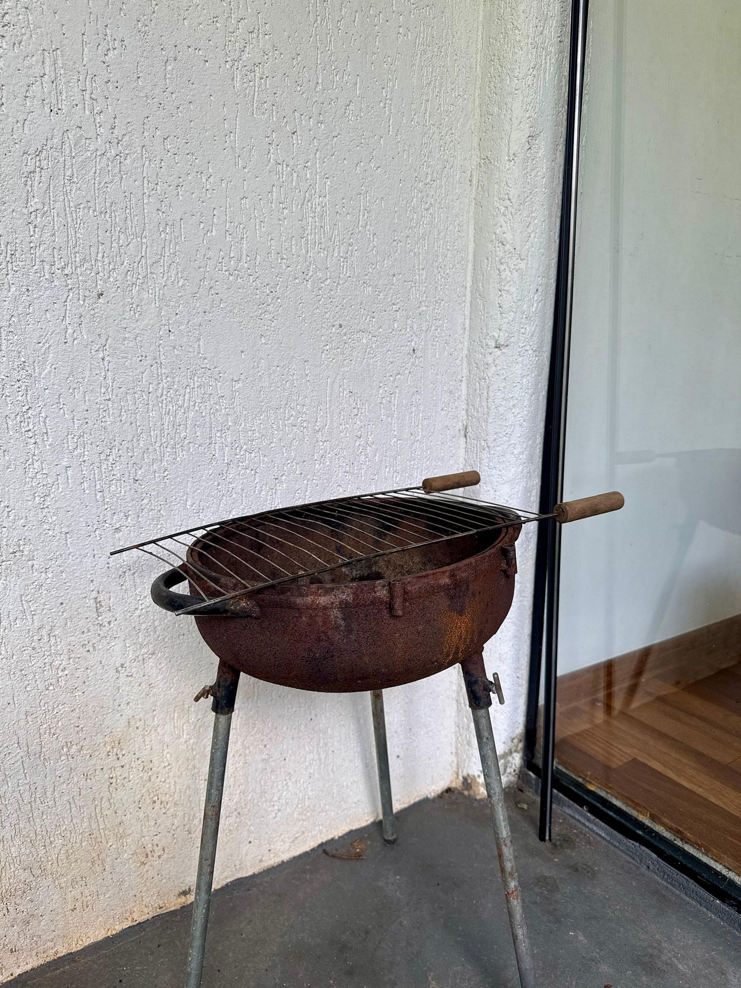 A portable barbecue grill with a metal grate and wooden handles, placed against a white wall with a glass door in the background.