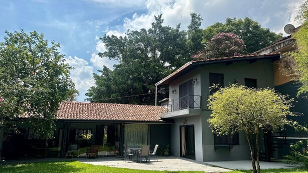 A two-story house with a tiled roof, a balcony, and a garden area with a lawn and a small tree. - Charme na Granja - Condomínio (Carapicuiba)