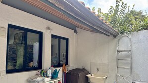 A small, enclosed outdoor area with a white tiled wall, a sink, a ladder, and a shelf with cleaning supplies. - Charme na Granja - Condomínio (Carapicuiba)