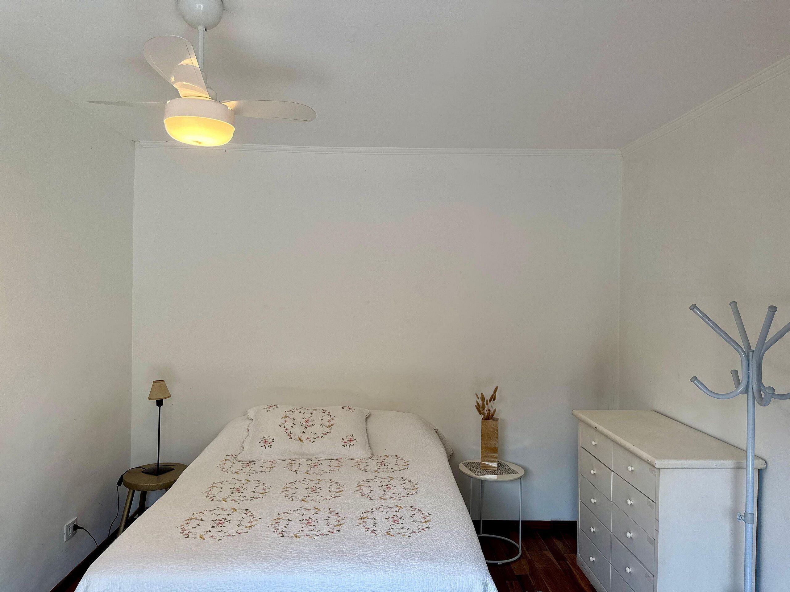 A bedroom with a bed, a ceiling fan, a bedside table with a lamp, a dresser, and a coat rack.