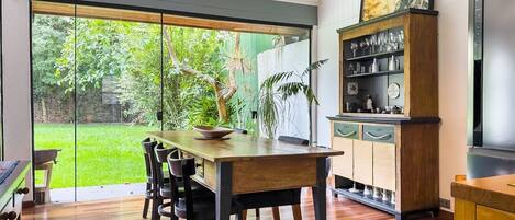 A dining area with a wooden table and chairs, a glass door leading to a garden, a wooden cabinet with glassware, and a television on the wall.