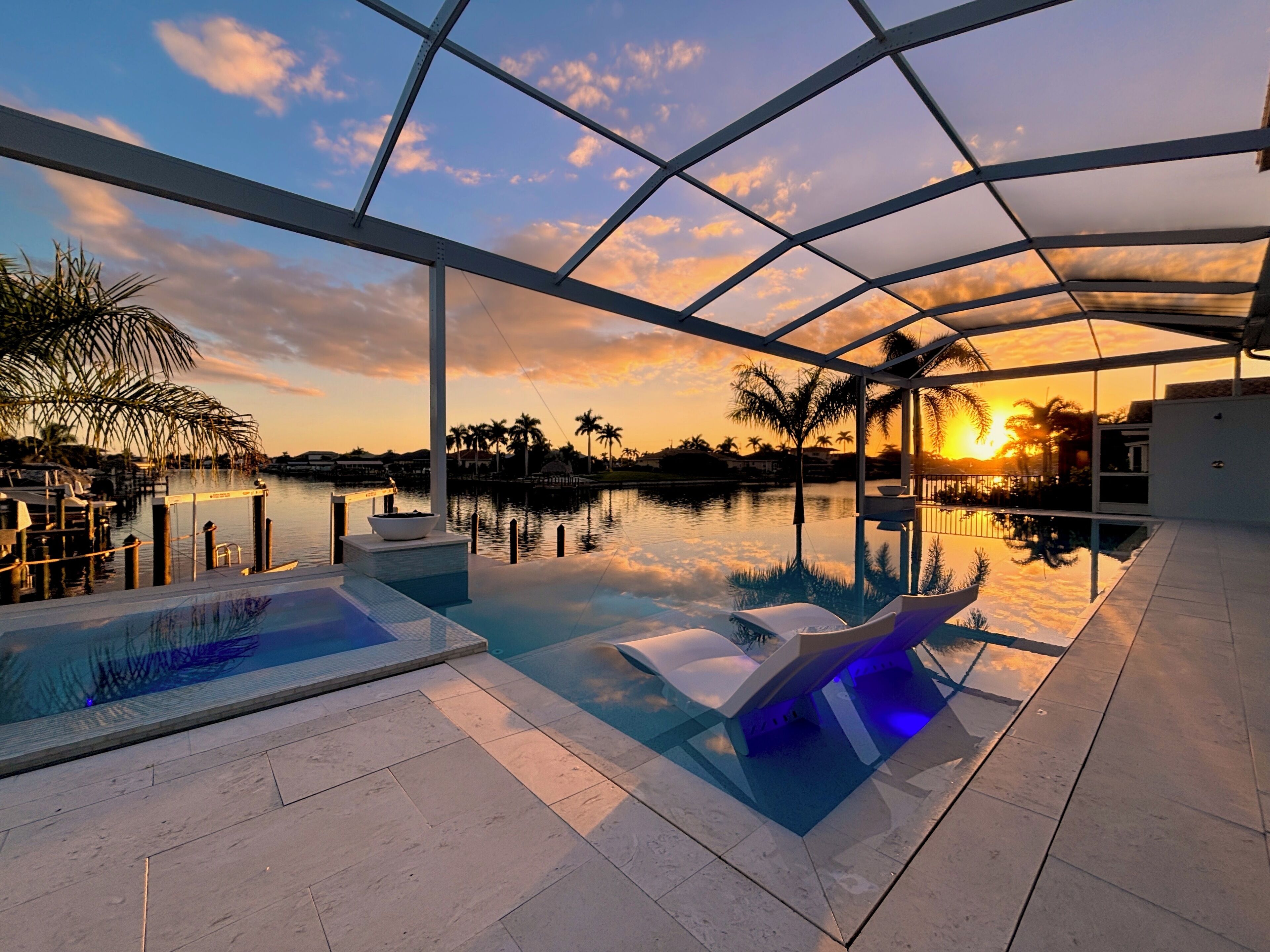 Ein Pool ohne Ende mit Blick auf einen Sonnenuntergang über dem Wasser, umgeben von Palmen und einem modernen Bauwerk.