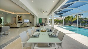 Un comedor moderno con vista a la piscina, un salón con televisor y una cocina al fondo.
