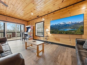 A wooden interior with a large window, a table, chairs, a sofa, and a wall-mounted TV displaying a mountain landscape.